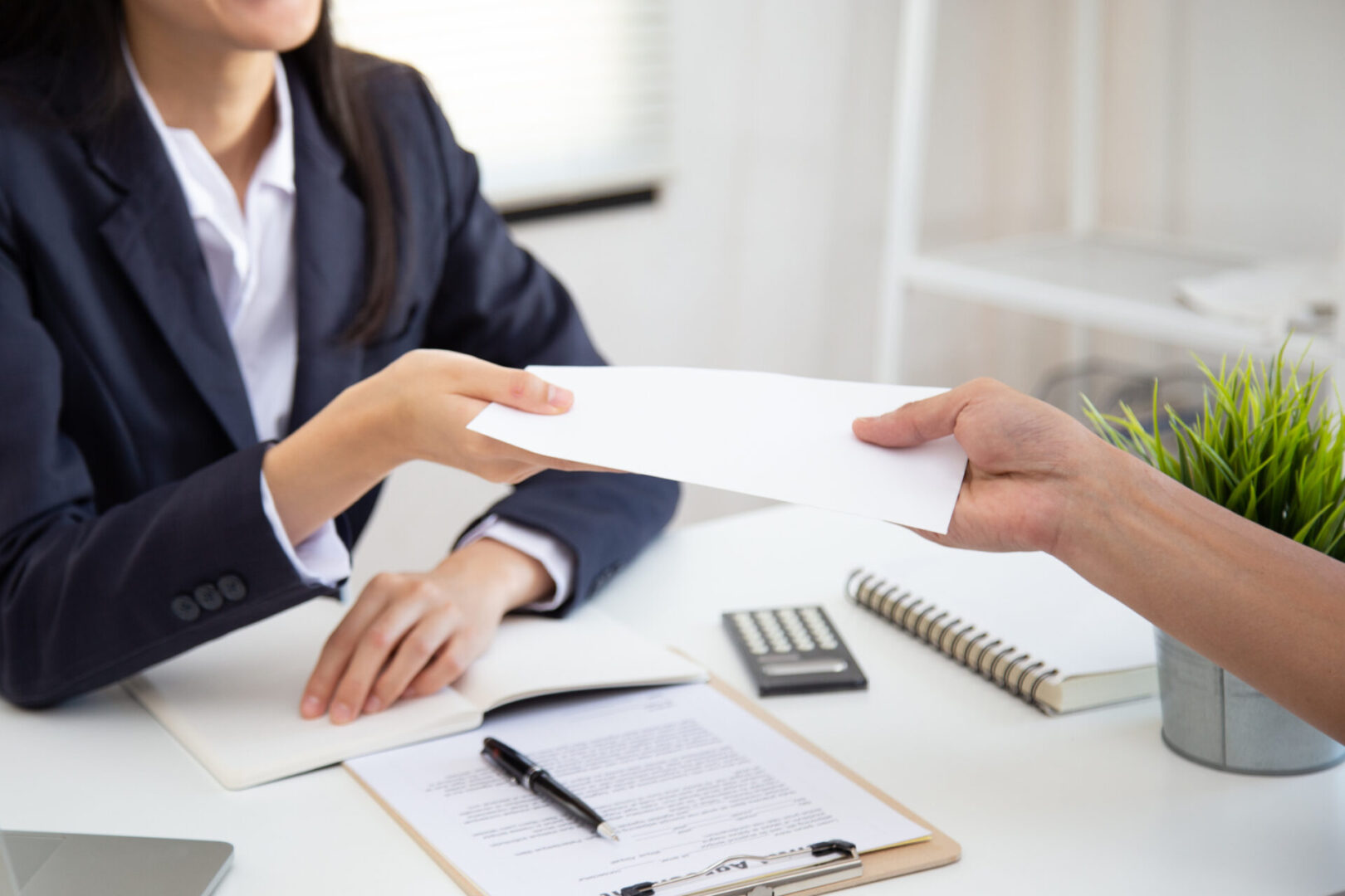 A person handing a document to another at a desk with papers and a calculator.