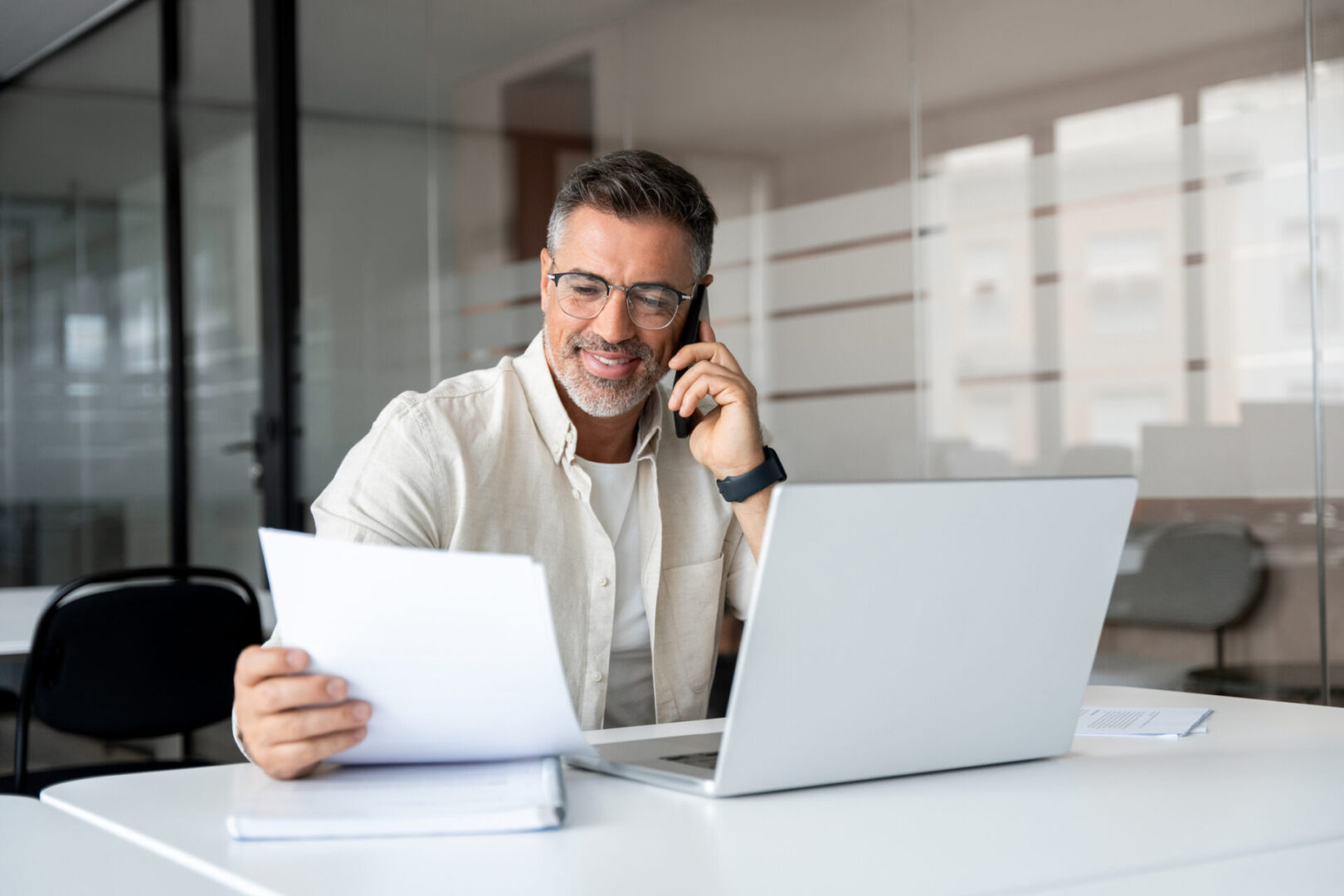 Man on phone reviewing documents with laptop in modern office.