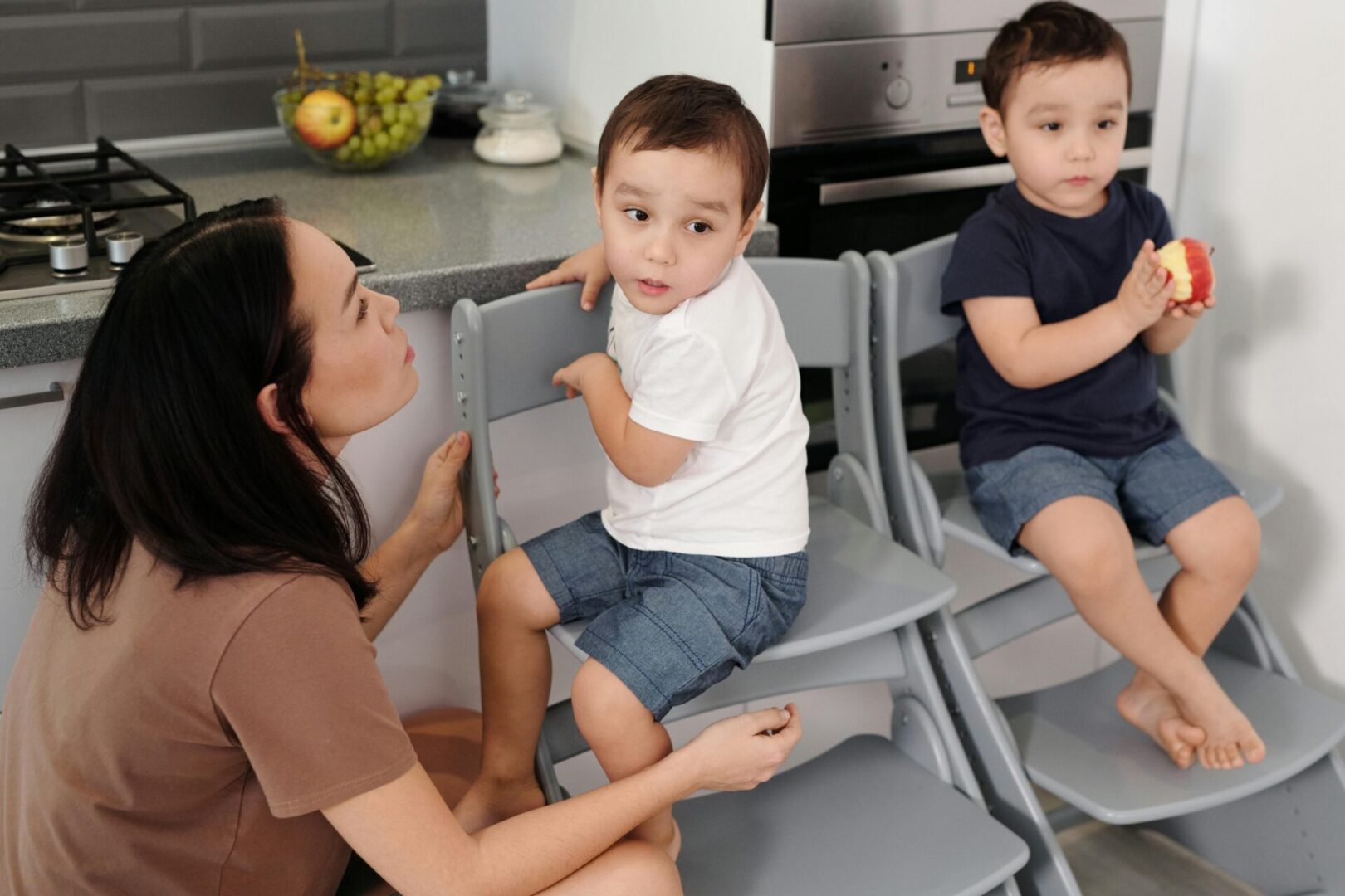 Mother kneeling beside two young boys sitting on chairs in a kitchen.