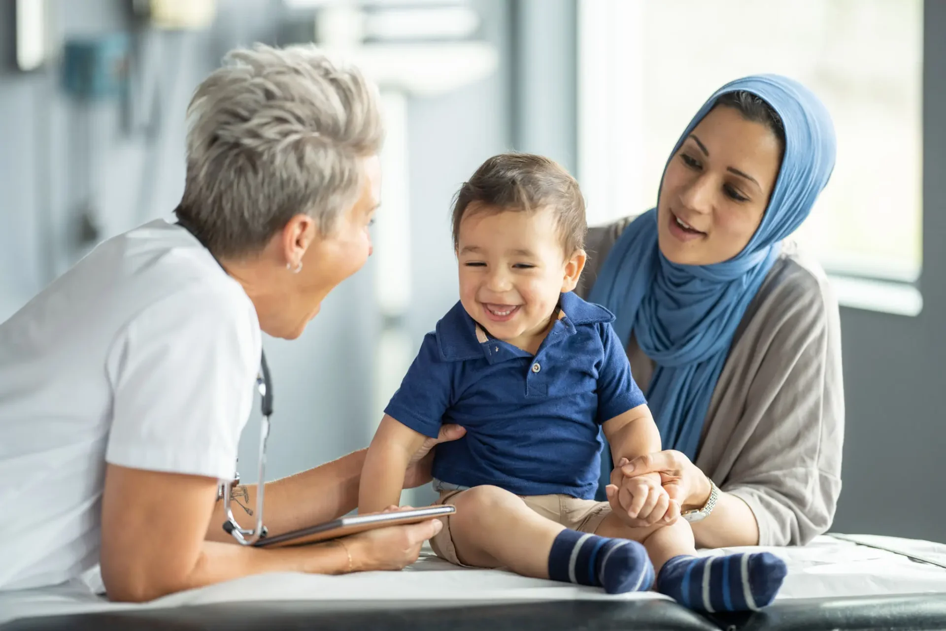A female doctor interacts with a smiling toddler and his mother in a clinic.