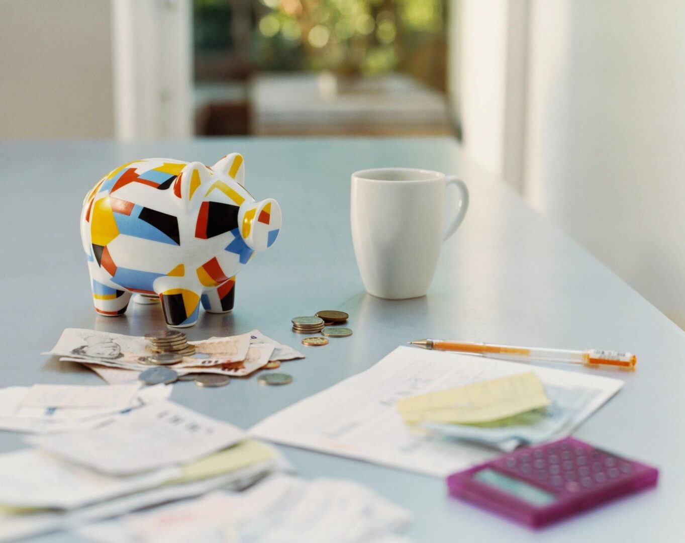 Colorful piggy bank, coins, and bills on a table with a coffee mug and calculator.